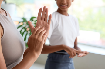Two people signing together indoors, with one person smiling in the background, representing communication and change through NZSL.
